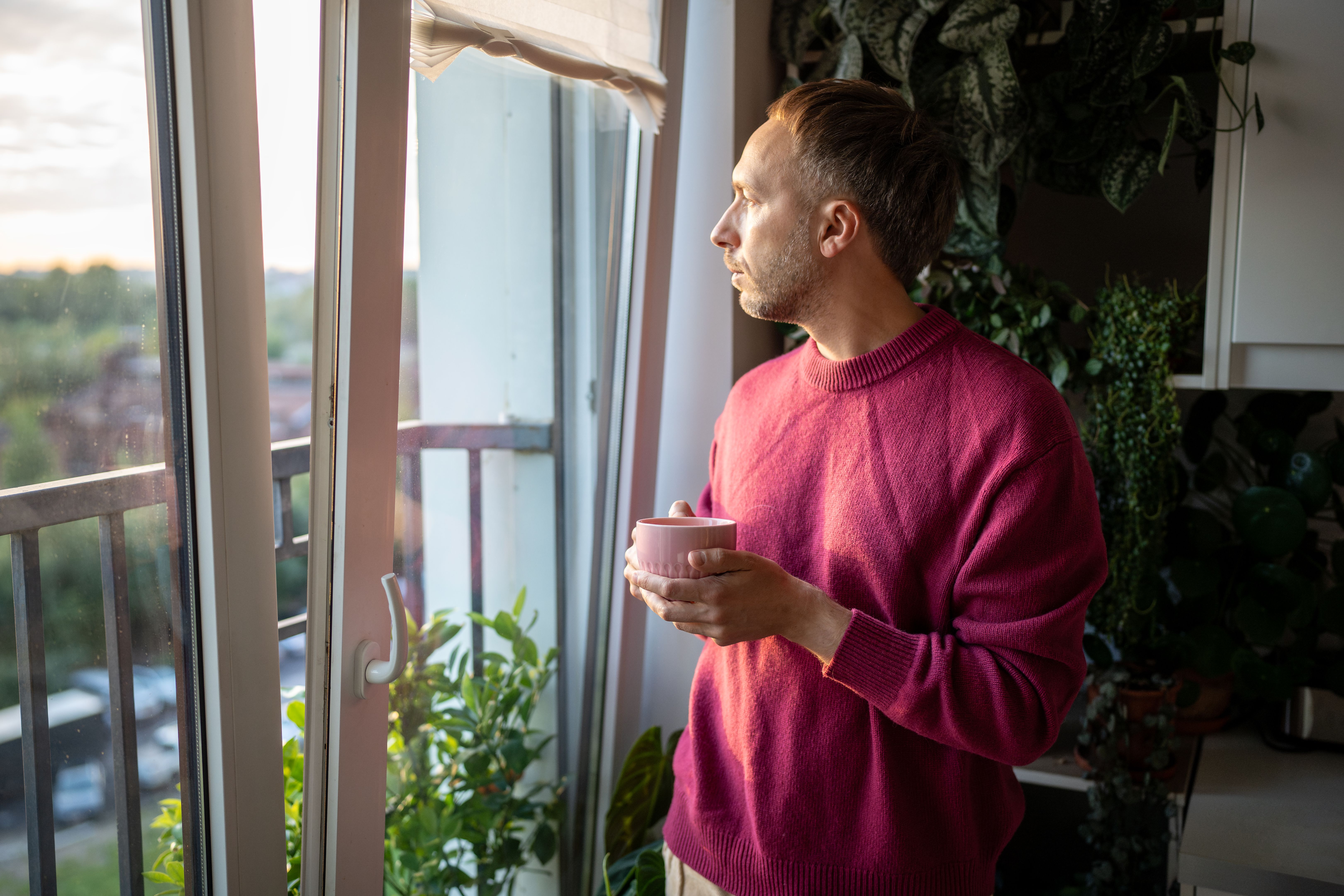 Pensive reflective man drinking tea, gazing out window alone in quiet somber mood near balcony