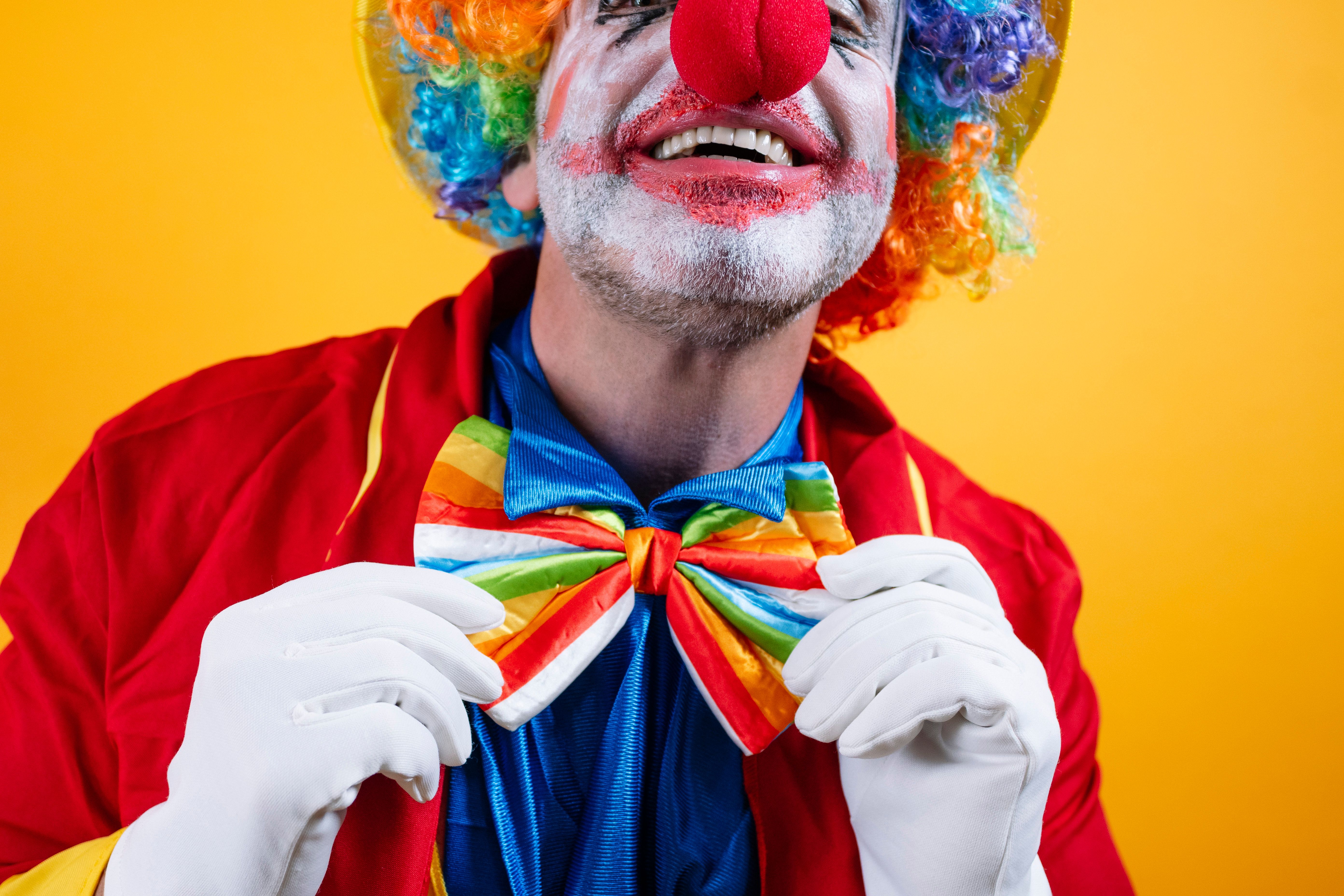 Happy clown adjusts rainbow bow tie, vibrant yellow background.