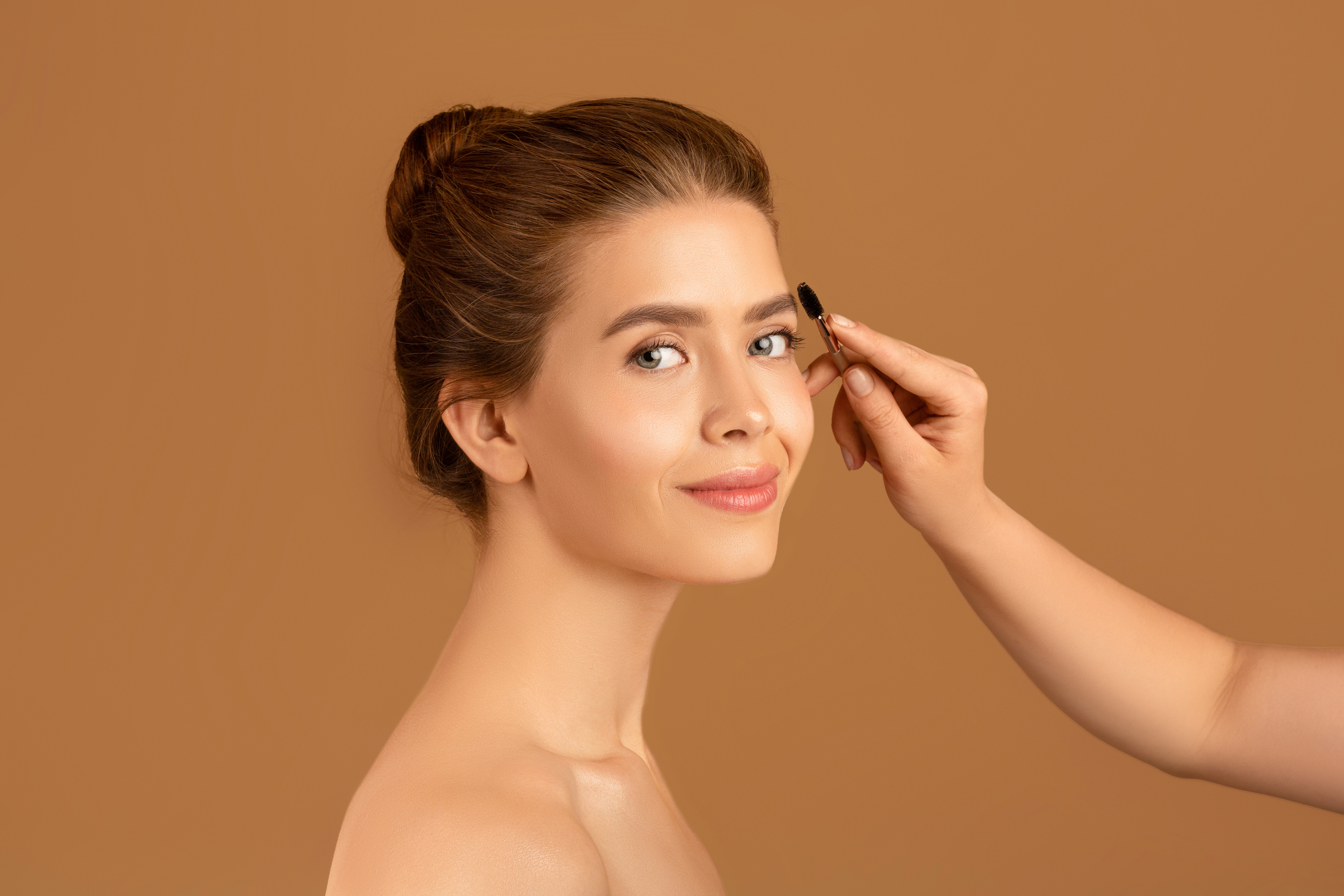 Makeup artist applying eyebrow mascara on millennial woman's face over brown studio background. Wellness and beauty