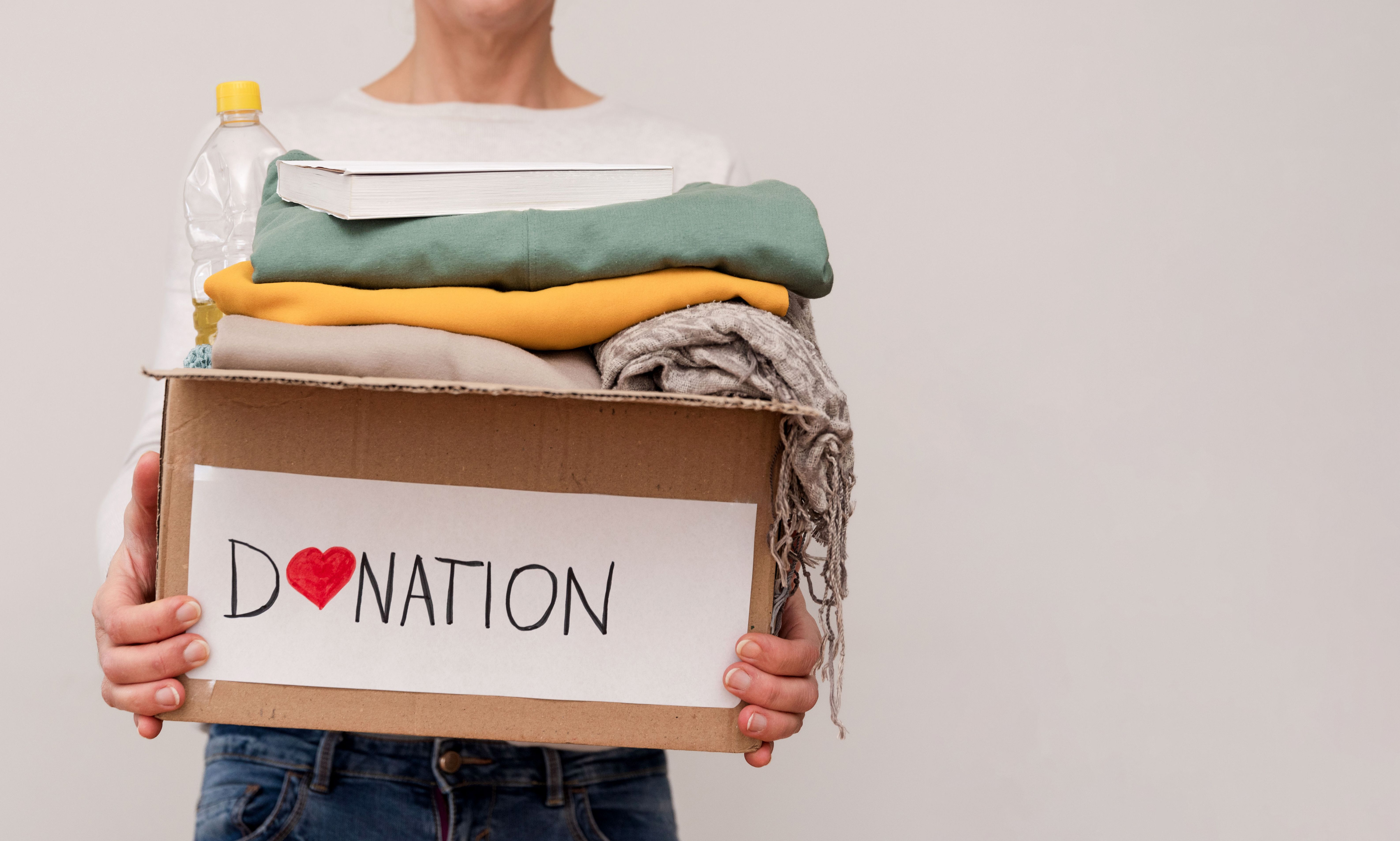 Close up of Volunteer holding donation box with clothes, food and book on grey background with copy space