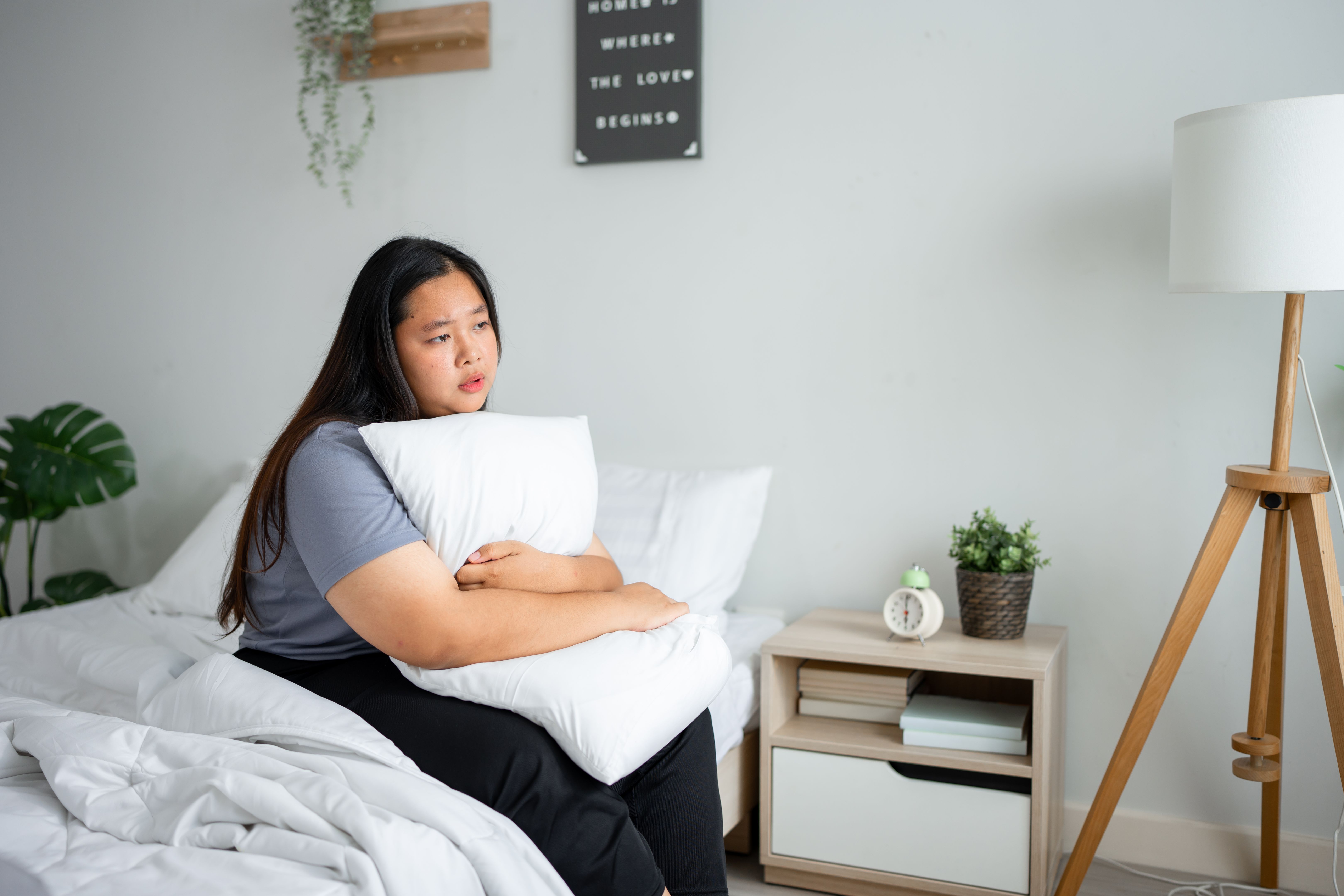 Asian depressed plus-size woman sitting alone on bed in bedroom at home. Attractive young overweight female feeling sad, lonely and upset with life problem and health care in quiet room in house.