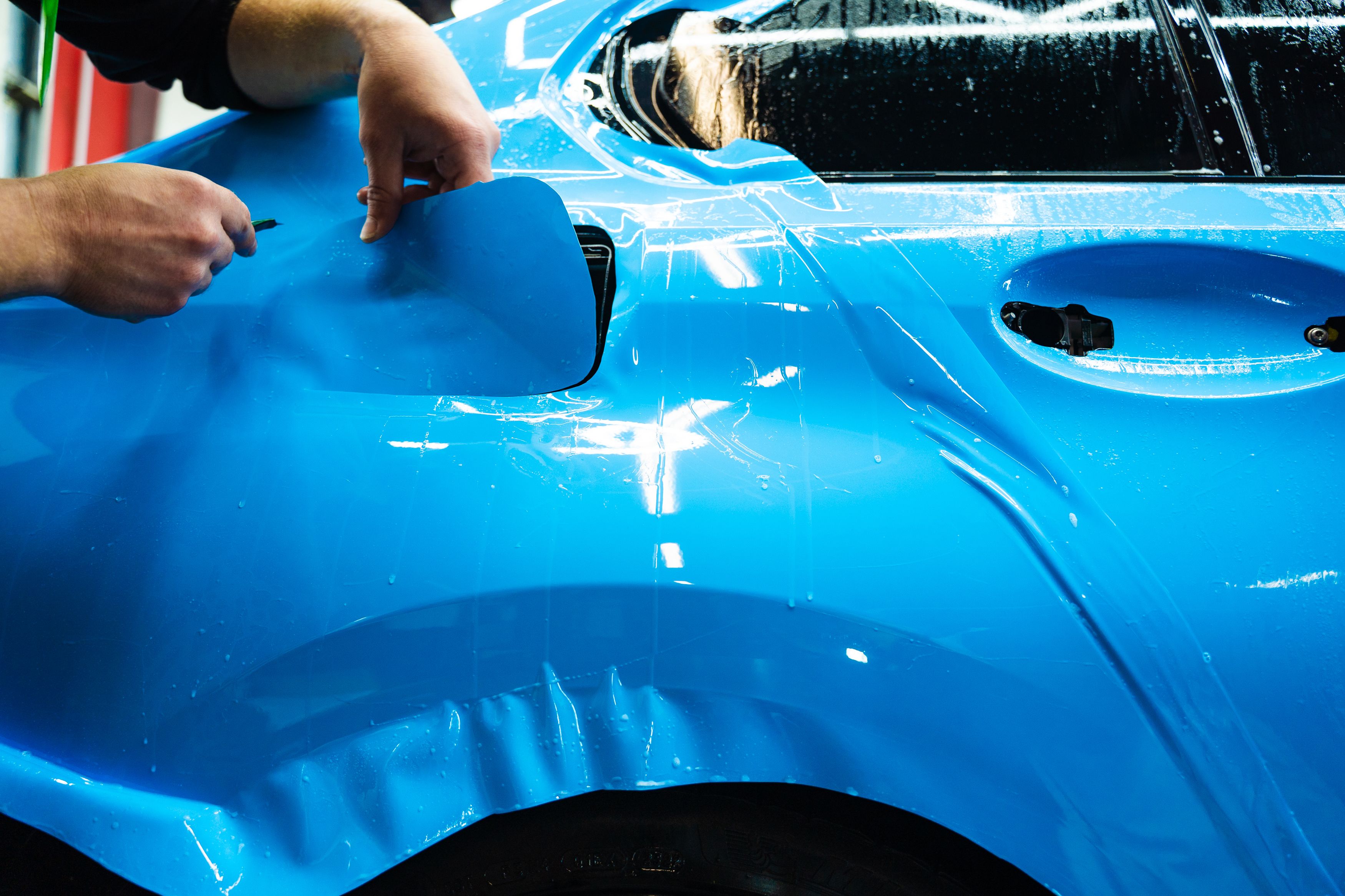 Detailing technician trims blue PPF on a car panel for perfect contour fit. Cutting blue PPF with blade. Cutting blue paint protection film with blade during detailing job ensures precision edges.