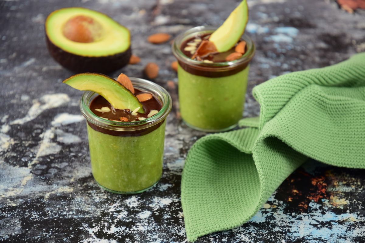 Avocado pudding with chocolate sauce and chopped almond nut in glass jars on grey background