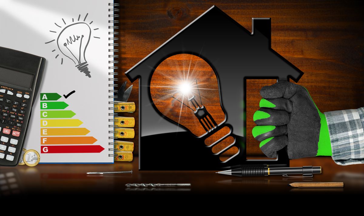 Energy Efficiency - Hand with work glove holding a model house (3D illustration) with energy efficiency rating and a light bulb. On a desk with reflections