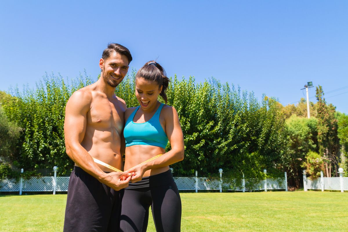 Delighted young athletic couple in sportswear measuring their waistline after a workout at the park. Healthy lifestyle and fitness concept.