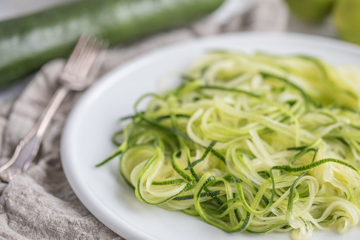 A white plate with zucchini noodles, zoodles, fresh pasta made of green squash. The food is  on a white plate, with a linen cloth and a fork  next to it.